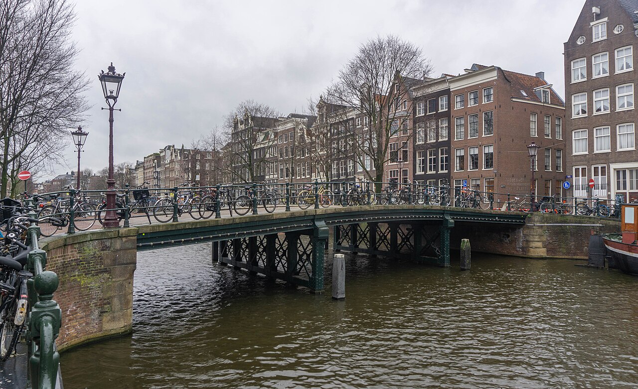 Bicycles chained along a canal bridge railing, with a boat passing underneath