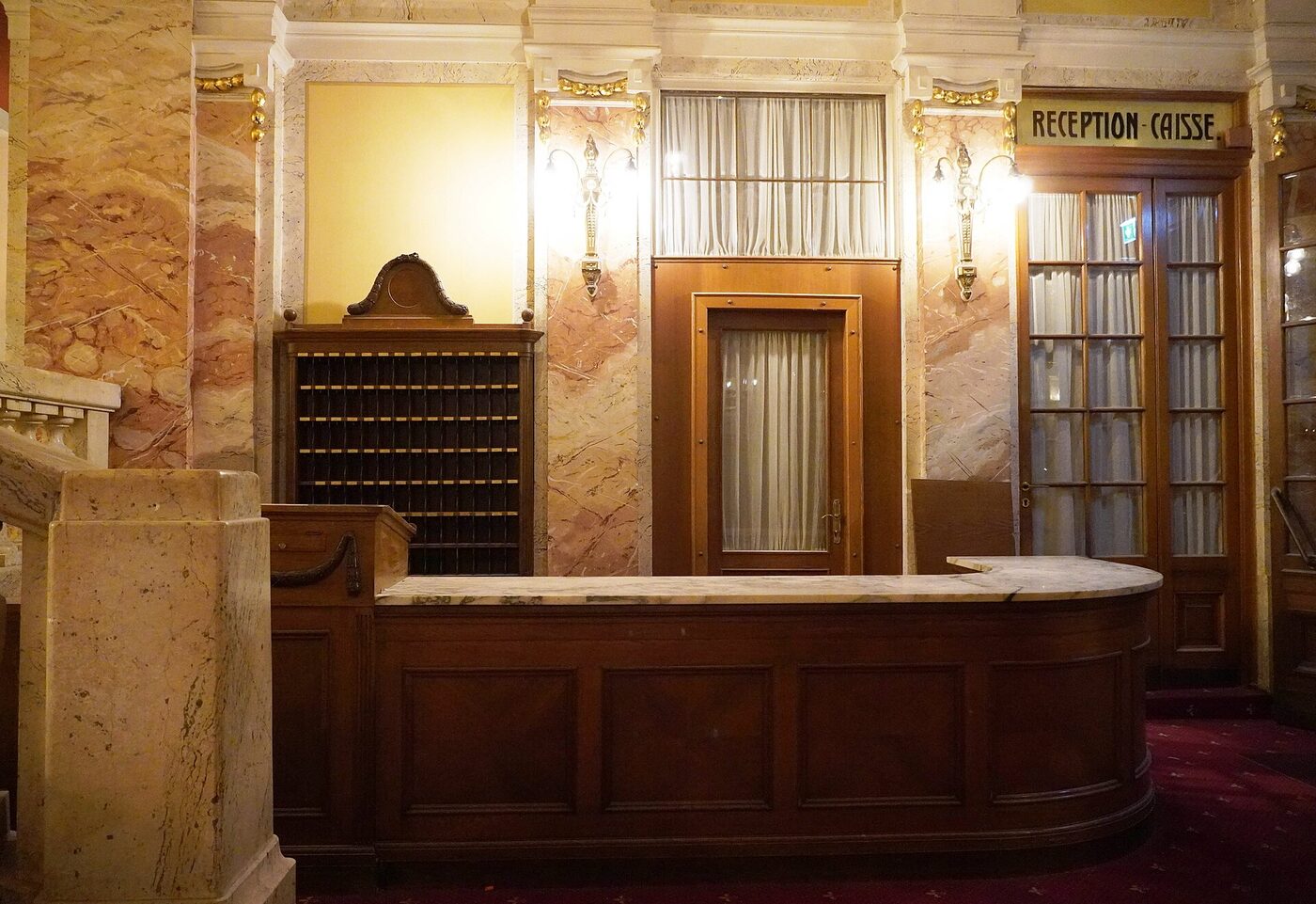 The reception desk of a grand hotel, with a wall of numbered key pigeonholes behind it