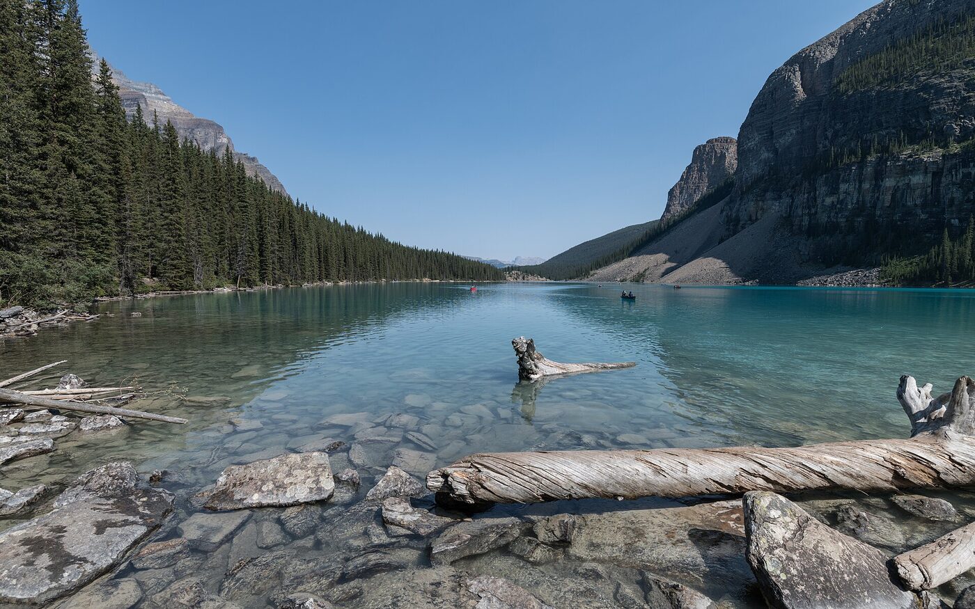 A turquoise glacial lake ringed by forested slopes and rock walls in the Canadian Rockies, with canoes on the far water