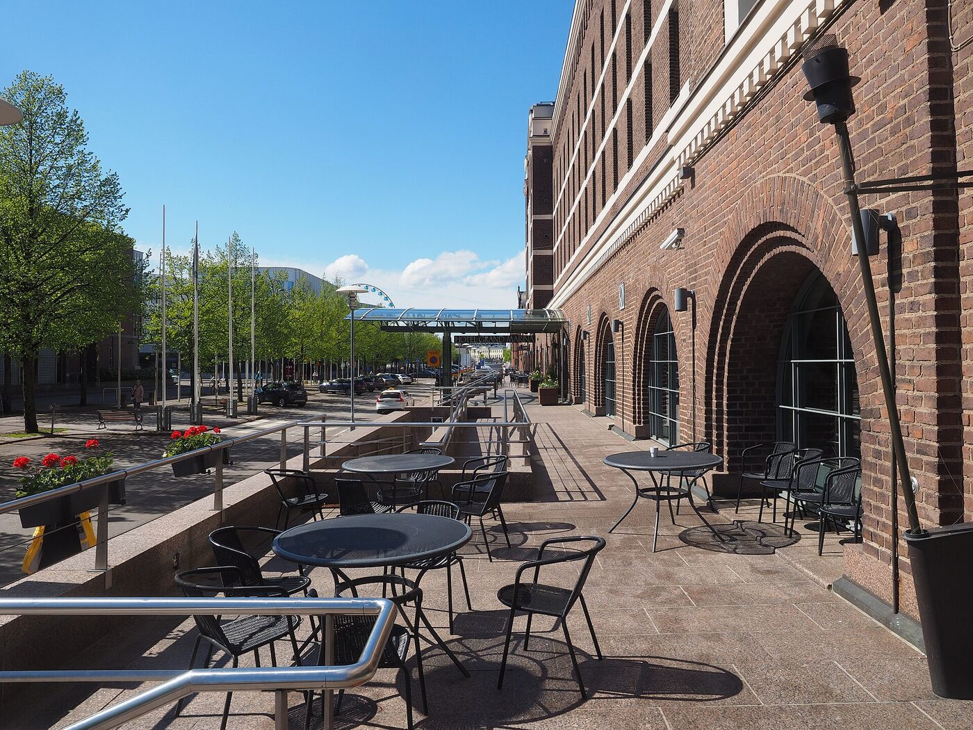 Empty tables and chairs on a hotel terrace beside a brick facade on a sunny city street