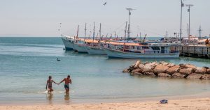 Two people wading in shallow sea water beside moored fishing boats